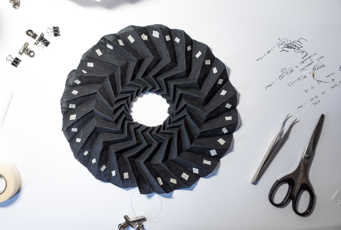 A black origami-pleated fabric ring on a workbench with tools and calculations